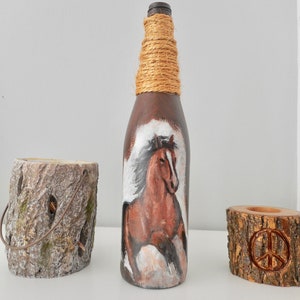 May include: A brown glass bottle with a painted image of a brown horse running. The bottle is wrapped with natural twine around the neck. The bottle is sitting on a white surface.