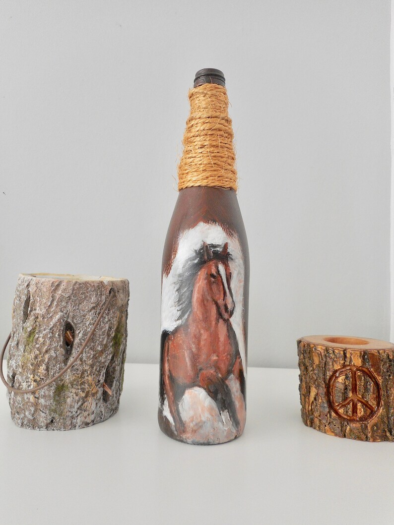 May include: A brown glass bottle with a painted image of a brown horse running. The bottle is wrapped with natural twine around the neck. The bottle is sitting on a white surface.
