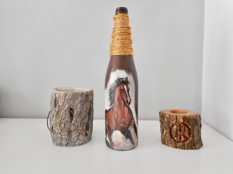 May include: A brown glass bottle with a horse painted on it, wrapped in twine. The bottle is next to two wooden candle holders, one with a peace sign carved into it.