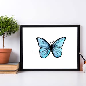 May include: A framed print featuring a watercolor illustration of a blue butterfly with black outlines. The artwork is displayed on a white surface, alongside a small potted plant, books, and decorative items.