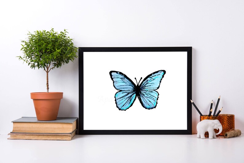 May include: A framed print featuring a watercolor illustration of a blue butterfly with black outlines. The artwork is displayed on a white surface, alongside a small potted plant, books, and decorative items.