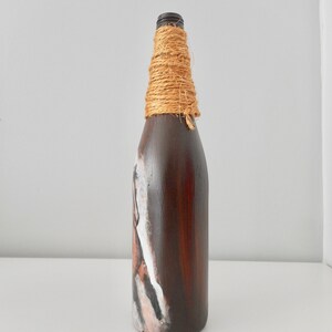 May include: A brown glass bottle with a white and brown painted design. The bottle is wrapped with natural twine around the neck.