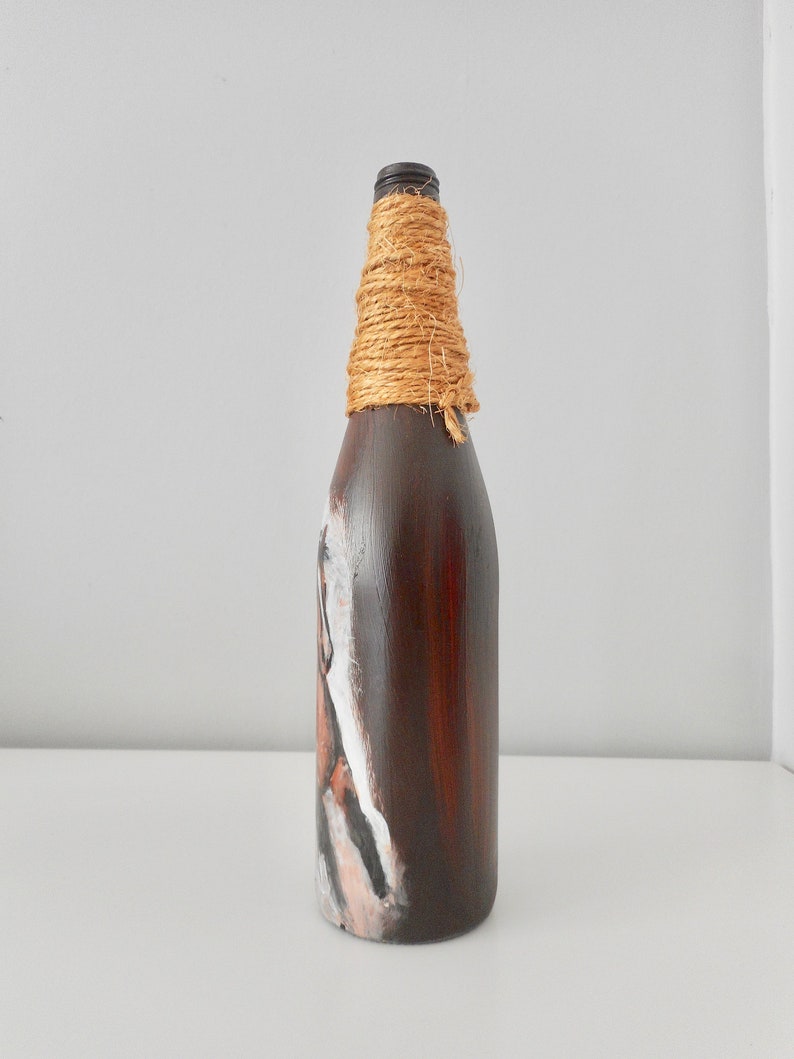 May include: A brown glass bottle with a white and brown painted design. The bottle is wrapped with natural twine around the neck.