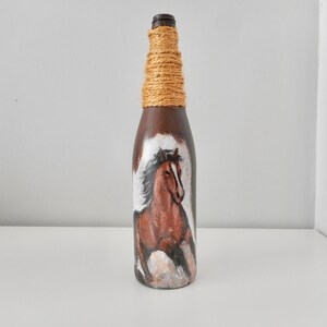 May include: A brown glass bottle with a painted image of a brown horse with a white mane and tail. The bottle is wrapped with twine around the neck.