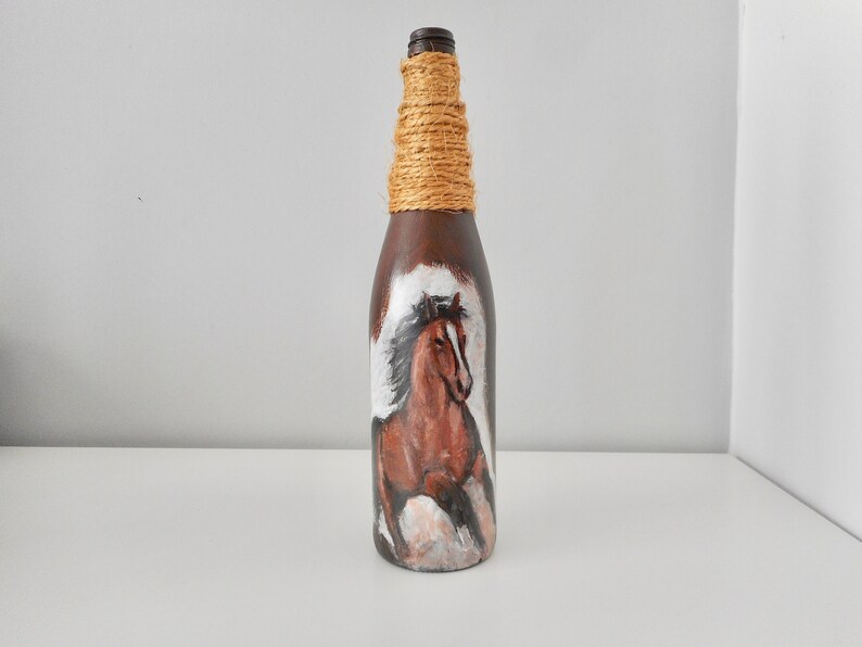 May include: A brown glass bottle with a painted image of a brown horse with a white mane and tail. The bottle is wrapped with twine around the neck.