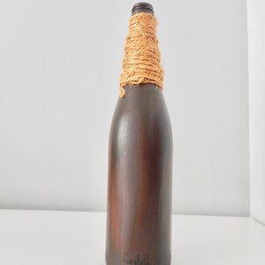 May include: A dark brown glass bottle with a natural fiber rope wrapped around the neck. The bottle is painted with a wood grain pattern and has the text "Hersbeth 14" written on the bottom.