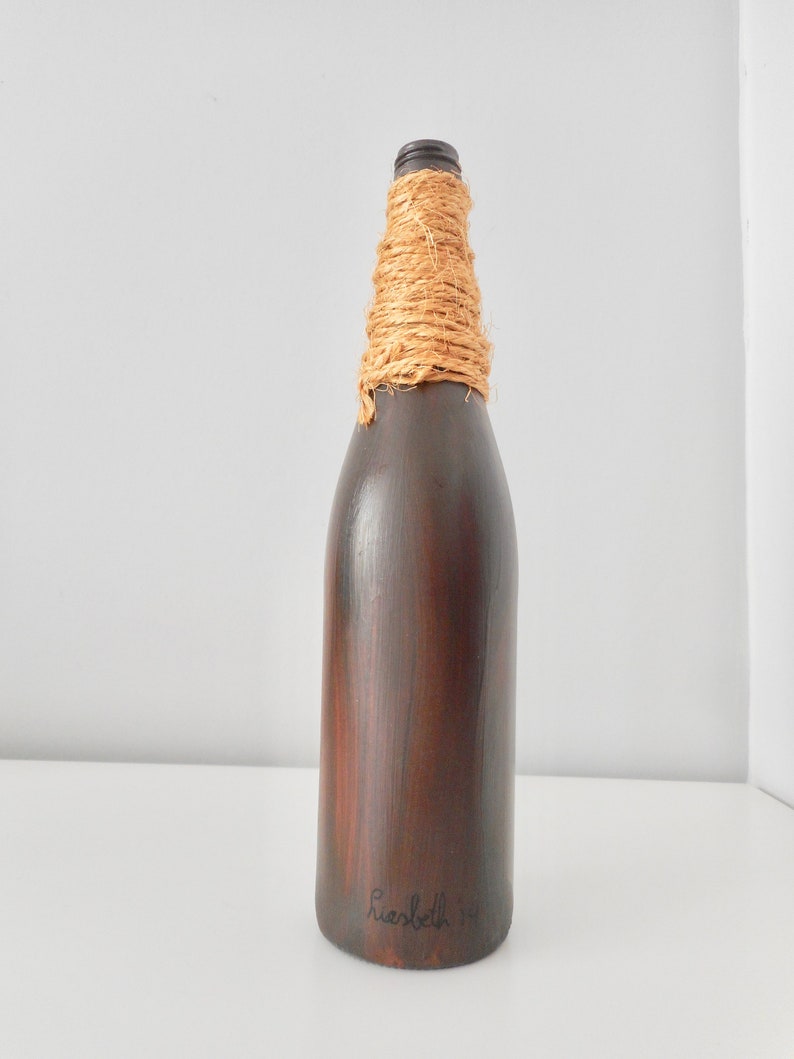 May include: A dark brown glass bottle with a natural fiber rope wrapped around the neck. The bottle is painted with a wood grain pattern and has the text "Hersbeth 14" written on the bottom.