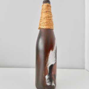 May include: A brown glass bottle with a white and brown painted design. The bottle is wrapped with natural twine around the neck.