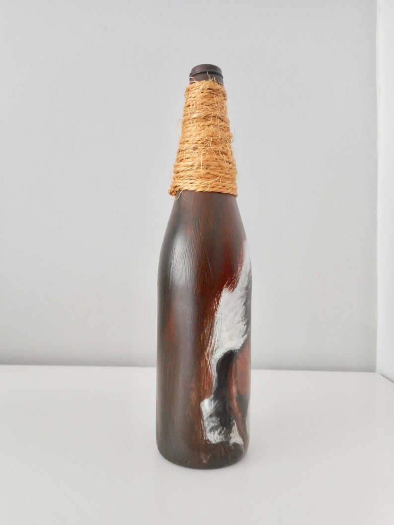 May include: A brown glass bottle with a white and brown painted design. The bottle is wrapped with natural twine around the neck.