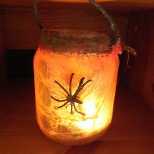 Op de afbeelding: Een glazen pot met een spinnenweb-ontwerp en een zwarte spin erin. De pot wordt van binnenuit verlicht met een warme gloed, wat een griezelige Halloween-sfeer creëert.
