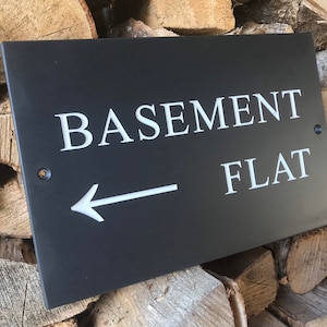 May include: A rectangular black slate sign featuring the white engraved text BASEMENT FLAT and a left-pointing arrow. The sign includes two pre-drilled holes for mounting, one on the left side and one on the right side, positioned near the edges.
