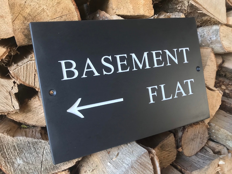 May include: A rectangular black slate sign featuring the white engraved text BASEMENT FLAT and a left-pointing arrow. The sign includes two pre-drilled holes for mounting, one on the left side and one on the right side, positioned near the edges.