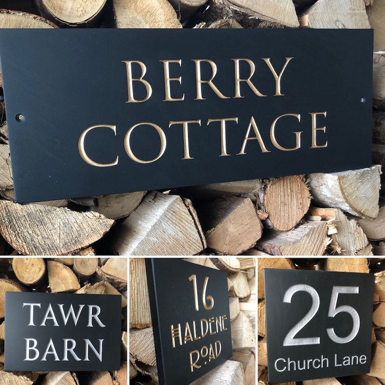 May include: A rectangular black house sign featuring engraved text in gold or white. The signs display various names and numbers including BERRY COTTAGE, TAWR BARN, 16 HALDENE ROAD, and 25 Church Lane. Each sign has small holes for mounting.