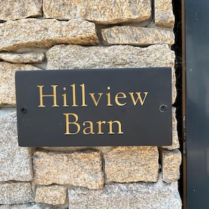 May include: A rectangular black slate sign with the text Hillview Barn engraved in gold lettering. The sign is mounted with two visible screws on a textured stone wall surface, featuring a dark gray frame edge on the right side of the frame.