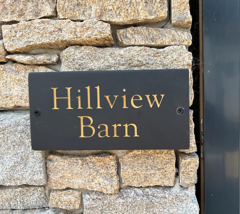 May include: A rectangular black slate sign with the text Hillview Barn engraved in gold lettering. The sign is mounted with two visible screws on a textured stone wall surface, featuring a dark gray frame edge on the right side of the frame.