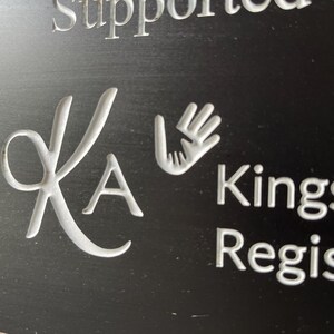 May include: A black sign featuring white engraved text and graphics. The design includes a stylized KA monogram, a graphic of two hands reaching toward each other, and the partial text "Supported by", "Kingsh", and "Registe" in a clean, sans-serif font.