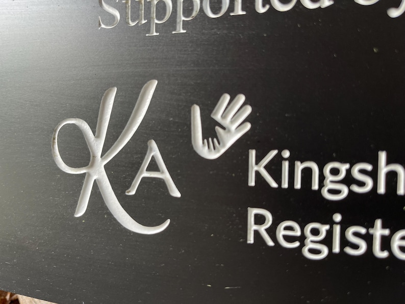 May include: A black sign featuring white engraved text and graphics. The design includes a stylized KA monogram, a graphic of two hands reaching toward each other, and the partial text "Supported by", "Kingsh", and "Registe" in a clean, sans-serif font.