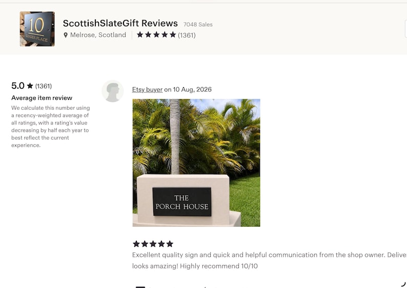 May include: A rectangular black slate sign with white engraved lettering that reads THE PORCH HOUSE. The sign is mounted horizontally on a light-colored, smooth, rectangular stone base positioned outdoors in front of green palm trees.