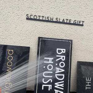 May include: Three rectangular black slate signs with engraved text. One sign reads TANGLEWOOD in gold, another reads BROADWAY HOUSE in white, and the third reads THE. A stream of water sprays across the center sign, creating a wet, glossy appearance.
