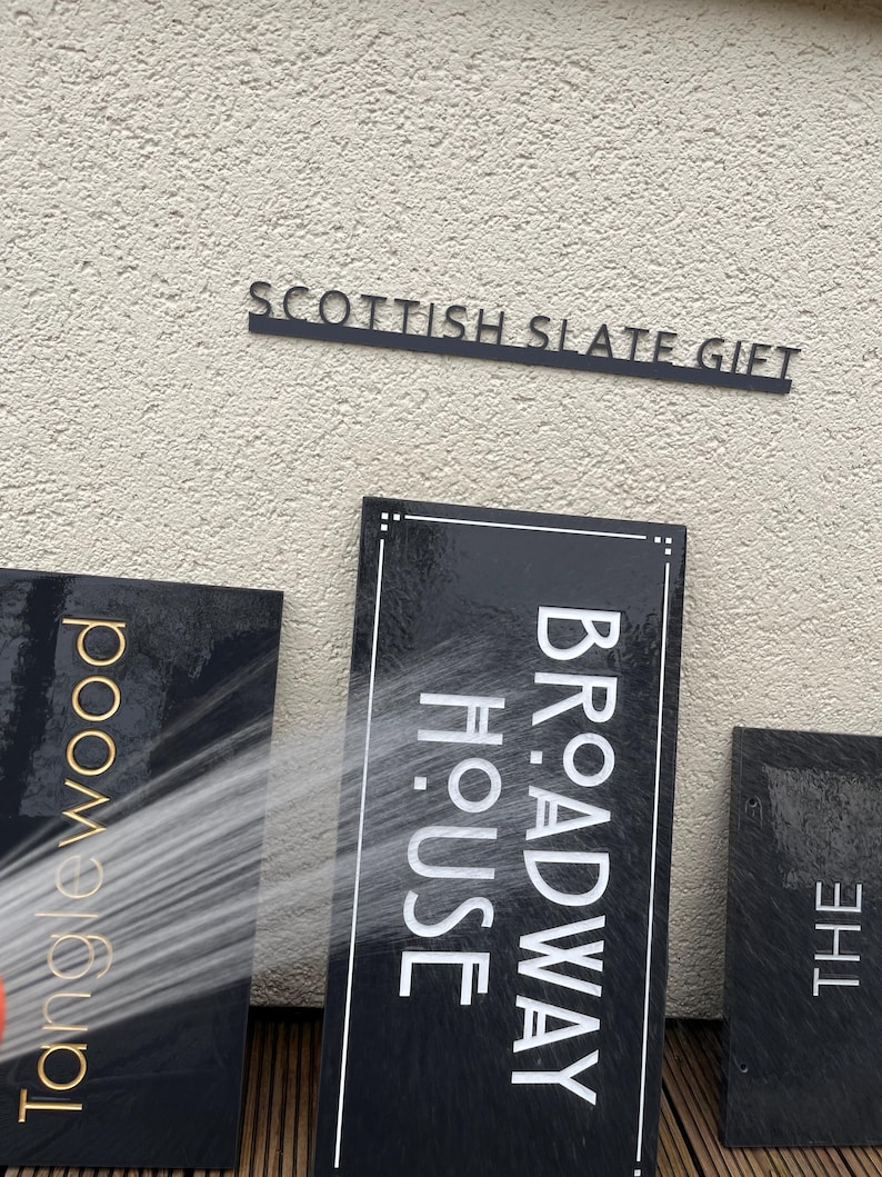 May include: Three rectangular black slate signs with engraved text. One sign reads TANGLEWOOD in gold, another reads BROADWAY HOUSE in white, and the third reads THE. A stream of water sprays across the center sign, creating a wet, glossy appearance.