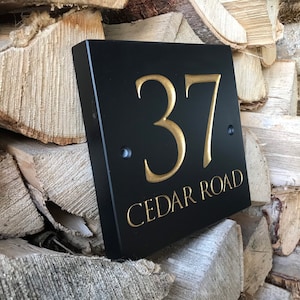 May include: A square black house sign featuring the number 37 and the text CEDAR ROAD in gold lettering. The sign has two visible mounting holes on the left and right sides, positioned centrally on the black surface with a smooth finish.