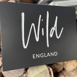 May include: A rectangular matte black sign featuring the word Wild in a large white cursive script font, with the word ENGLAND printed below in a smaller, clean white sans-serif typeface. The sign has a smooth, flat surface finish.