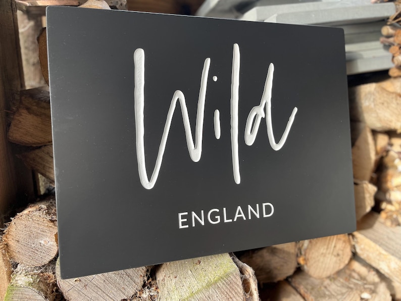 May include: A rectangular matte black sign featuring the word Wild in a large white cursive script font, with the word ENGLAND printed below in a smaller, clean white sans-serif typeface. The sign has a smooth, flat surface finish.