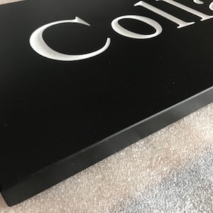 May include: A rectangular black sign featuring white, recessed, serif-style lettering that partially spells out the word Colla. The sign has a smooth, matte finish and a thick, solid edge, presented from an angled perspective to show depth.