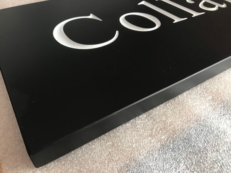 May include: A rectangular black sign featuring white, recessed, serif-style lettering that partially spells out the word Colla. The sign has a smooth, matte finish and a thick, solid edge, presented from an angled perspective to show depth.