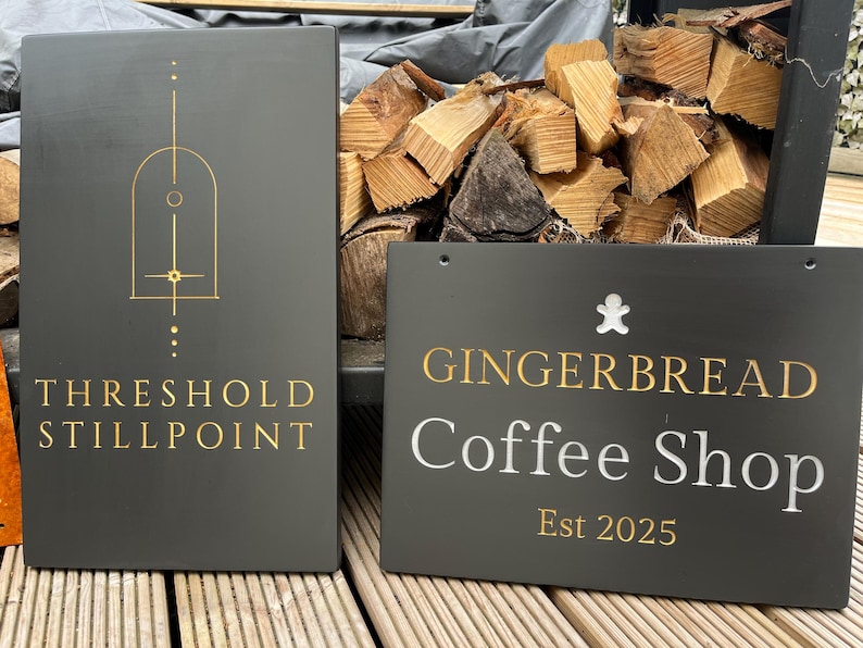 May include: Two rectangular dark gray signs. The left sign features a gold geometric arch design and the text THRESHOLD STILLPOINT. The right sign displays a white gingerbread man icon, the text GINGERBREAD Coffee Shop, and the gold text Est 2025.