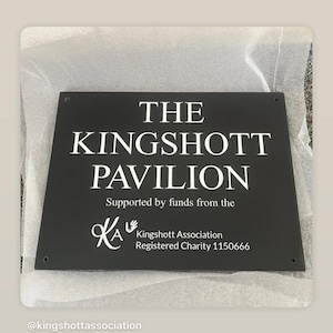 May include: A rectangular black sign with white text reading THE KINGSHOTT PAVILION, Supported by funds from the, KA Kingshott Association, Registered Charity 1150666. The sign features four small circular holes in the corners for mounting purposes.