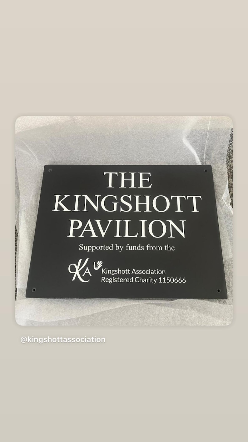 May include: A rectangular black sign with white text reading THE KINGSHOTT PAVILION, Supported by funds from the, KA Kingshott Association, Registered Charity 1150666. The sign features four small circular holes in the corners for mounting purposes.