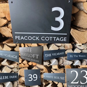 May include: A collection of rectangular dark gray slate house signs featuring white engraved text and numbers. The signs display various names such as Peacock Cottage, The Hayloft, The Vicarage, By The Waters, Glebe Farm, and The White House.