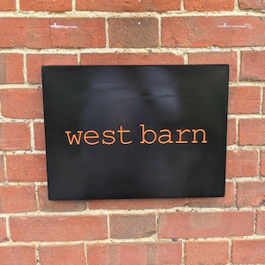 May include: A rectangular black sign with the text west barn written in orange lowercase letters. The sign has a smooth, matte finish and is mounted against a red brick wall with visible mortar lines between the rectangular bricks.