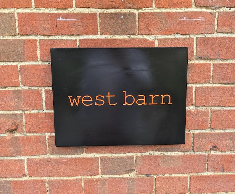 May include: A rectangular black sign with the text west barn written in orange lowercase letters. The sign has a smooth, matte finish and is mounted against a red brick wall with visible mortar lines between the rectangular bricks.