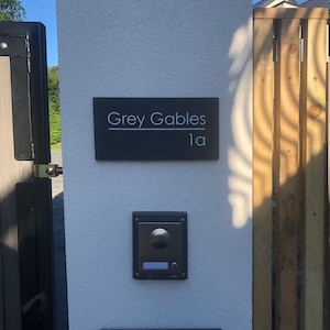 May include: A rectangular dark gray house sign with the white text Grey Gables and 1a, mounted on a white pillar above a silver and black intercom system. The sign features a horizontal line separating the house name from the number.