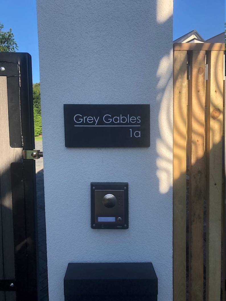 May include: A rectangular dark gray house sign with the white text Grey Gables and 1a, mounted on a white pillar above a silver and black intercom system. The sign features a horizontal line separating the house name from the number.