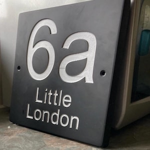 May include: A rectangular dark gray slate house sign featuring the engraved white text 6a and the words Little London below it. The sign has two circular mounting holes on the left and right sides, with a smooth, matte finish and clean edges.
