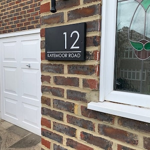 May include: A rectangular black house sign mounted on a brick wall. The sign features the white number 12 at the top, a horizontal white line, and the text KAYEMOOR ROAD in white capital letters at the bottom. The sign is secured with visible screws.