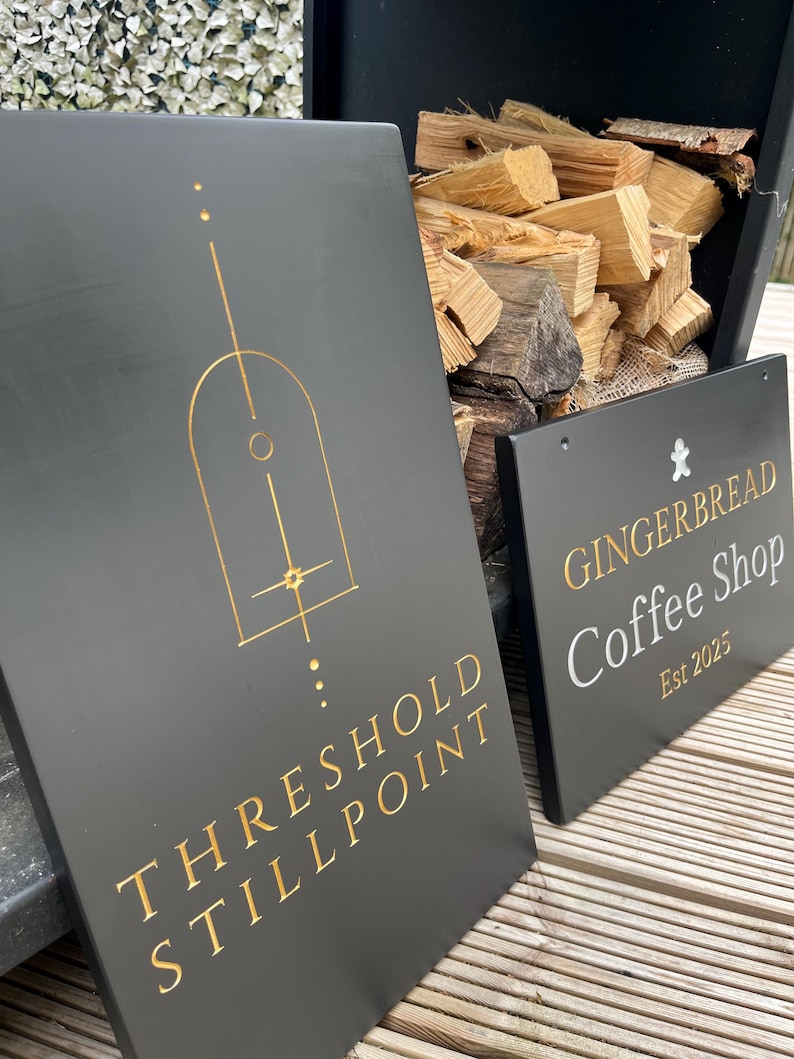 May include: Two rectangular black signs with gold lettering. The larger sign features a geometric arch design and the text "THRESHOLD STILLPOINT". The smaller sign displays a gingerbread man icon, the text "GINGERBREAD Coffee Shop", and "Est 2025".