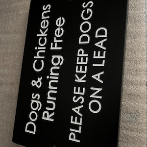 May include: A rectangular black sign with white engraved text that reads: Dogs & Chickens Running Free, PLEASE KEEP DOGS ON A LEAD. The sign features two small circular holes at the top and bottom center for mounting purposes.