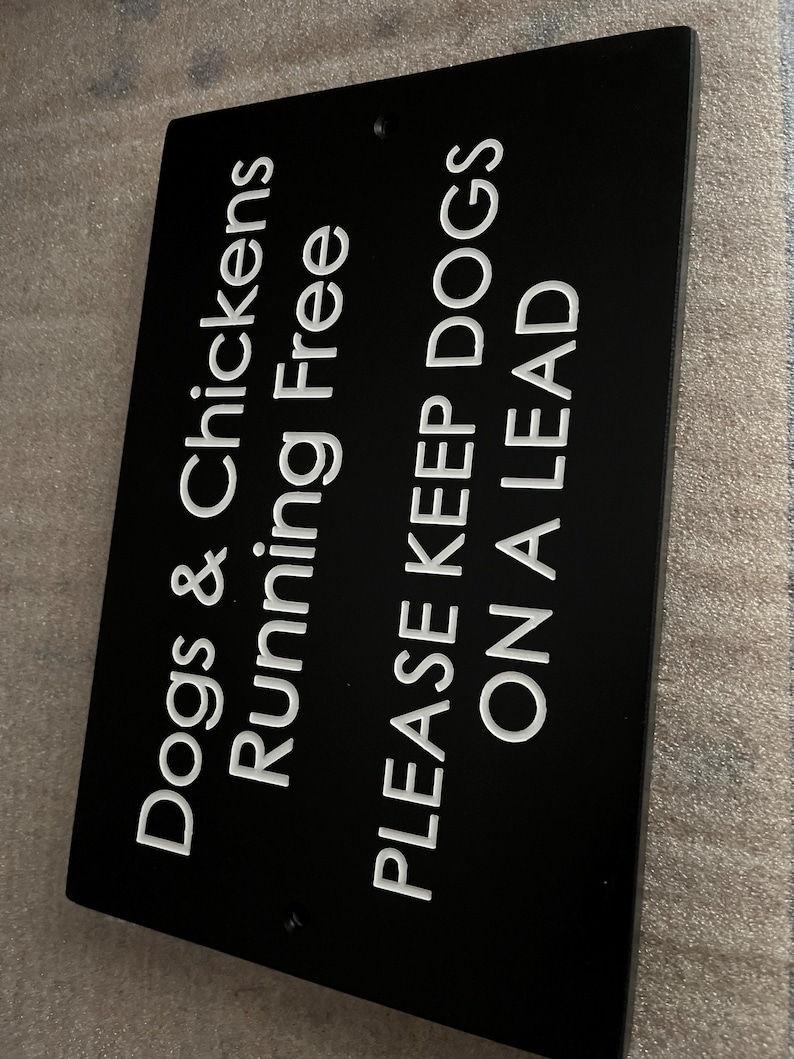 May include: A rectangular black sign with white engraved text that reads: Dogs & Chickens Running Free, PLEASE KEEP DOGS ON A LEAD. The sign features two small circular holes at the top and bottom center for mounting purposes.