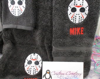 May include: Three dark grey towels with embroidered white hockey masks and the name "Mike" in red. The towels are on a wooden surface with a business card that says "Custom Creations by Cheryl" and a penguin illustration.
