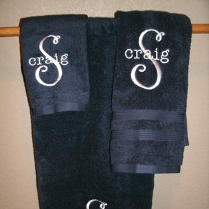 May include: Three dark blue towels with white embroidered lettering. The towels are hanging on a wooden rod. The lettering says "Craig" with a stylized letter "S".