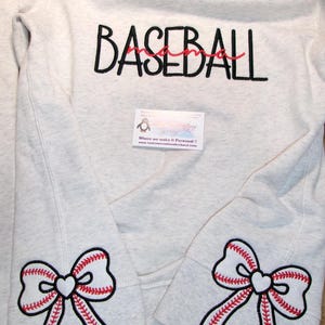 May include: Light gray sweatshirt with the word "BASEBALL" in black lettering and "mama" in red script. Baseball bow accents are embroidered on the sleeves. A business card is visible.
