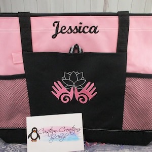 May include: Pink tote bag with black accents and a zipper closure. The bag has a large front pocket with a white lotus flower and two hands design embroidered on it. The bag is personalized with the name "Jessica".