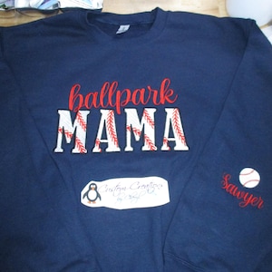 May include: Navy blue sweatshirt with the words "ballpark MAMA" in red and white, baseball-themed lettering. The name "Sawyer" is embroidered on the sleeve, along with a baseball graphic. A small penguin logo is visible.