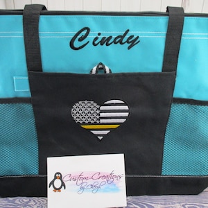 May include: A turquoise tote bag with black straps and a black zipper. The bag has a large front pocket with a black and white heart-shaped American flag with a yellow stripe. The bag is personalized with the name "Cindy" in black embroidery. The bag is sitting on a wooden surface.