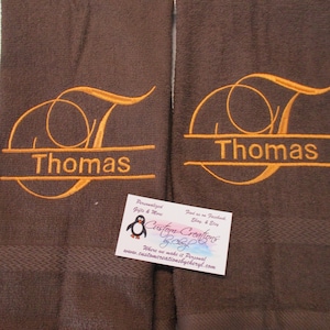 May include: Two brown towels with the name "Thomas" embroidered in orange on each towel. The towels have a textured surface and a white border. A business card for "Custom Creations by Cheryl" is in the foreground.