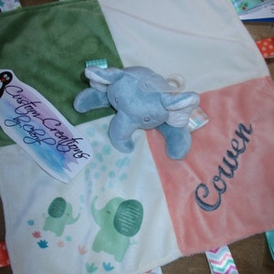 May include: A multi-colored baby blanket with a blue elephant plush toy. The blanket has a green, white, and pink patchwork design with various textures. The blanket has tags with the text "Custom Creations by Cheryl" and "Colton".